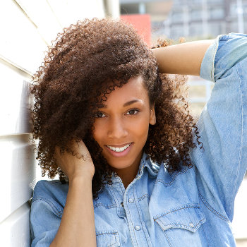 A smiling woman with curly hair wearing a denim shirt poses casually by a wall.