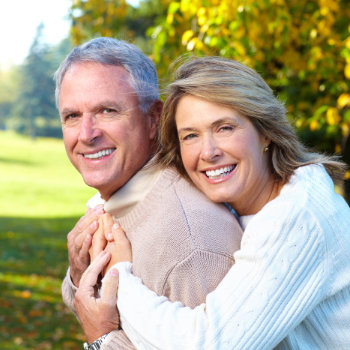 A smiling middle-aged couple embracing each other outdoors with trees in the background.