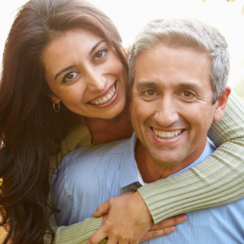 A smiling woman embracing a man from behind, both looking at the camera.