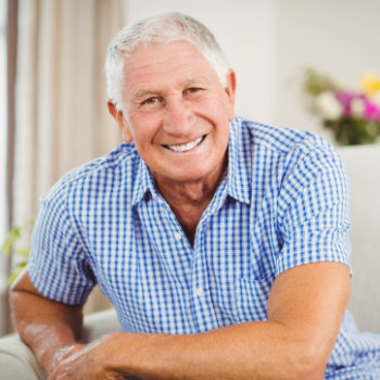 Smiling elderly man in a blue checked shirt sitting indoors.