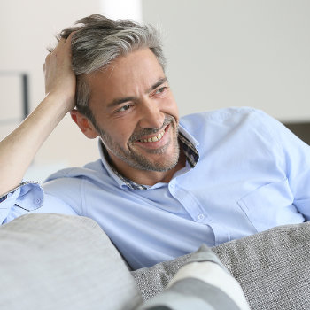 A smiling middle-aged man with gray hair, resting his hand on his head, reclining comfortably on a gray sofa.