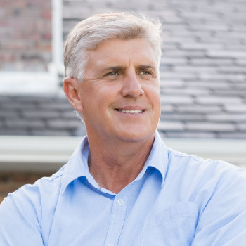 Mature man with gray hair smiling while wearing a blue shirt, with a blurred background.