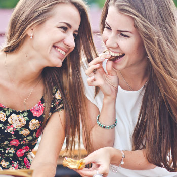 Two women smiling and sharing a chocolate bar outdoors.