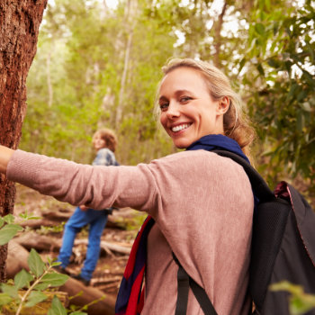 A woman with a backpack smiling at the camera while touching a tree, with a child in the background walking through the forest.