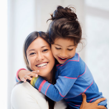 A smiling woman embracing a joyful young girl who is hugging her from behind.