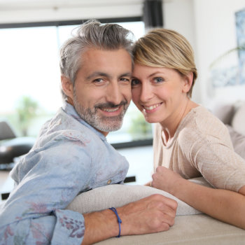 A smiling middle-aged couple posing closely together on a sofa in a bright living room.
