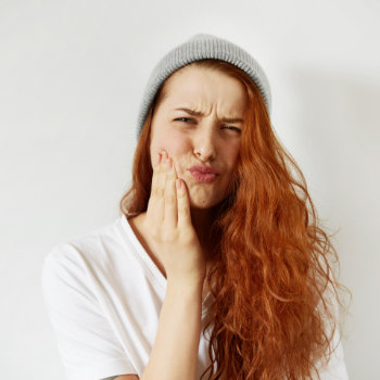 A young woman with red hair wearing a grey beanie touches her cheek and winces, as if she's experiencing pain or discomfort.