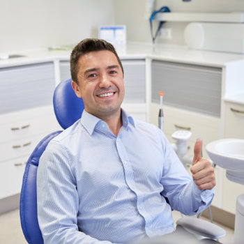 A man in a blue shirt sitting in a dental clinic office, giving a thumbs up.