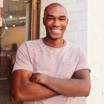 A smiling man with crossed arms wearing a striped shirt, standing by a brick wall.