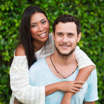 A smiling couple embracing in front of a leafy green hedge.