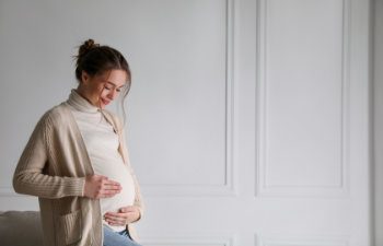 A pregnant woman stands indoors, looking down and gently holding her belly with both hands. She wears a beige cardigan over a white turtleneck.
