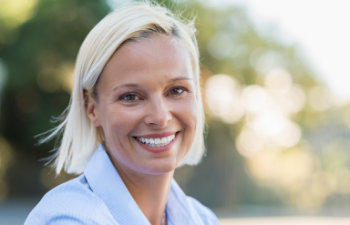 A woman with blonde hair and wearing a light blue shirt smiles outdoors with a blurred background of greenery.